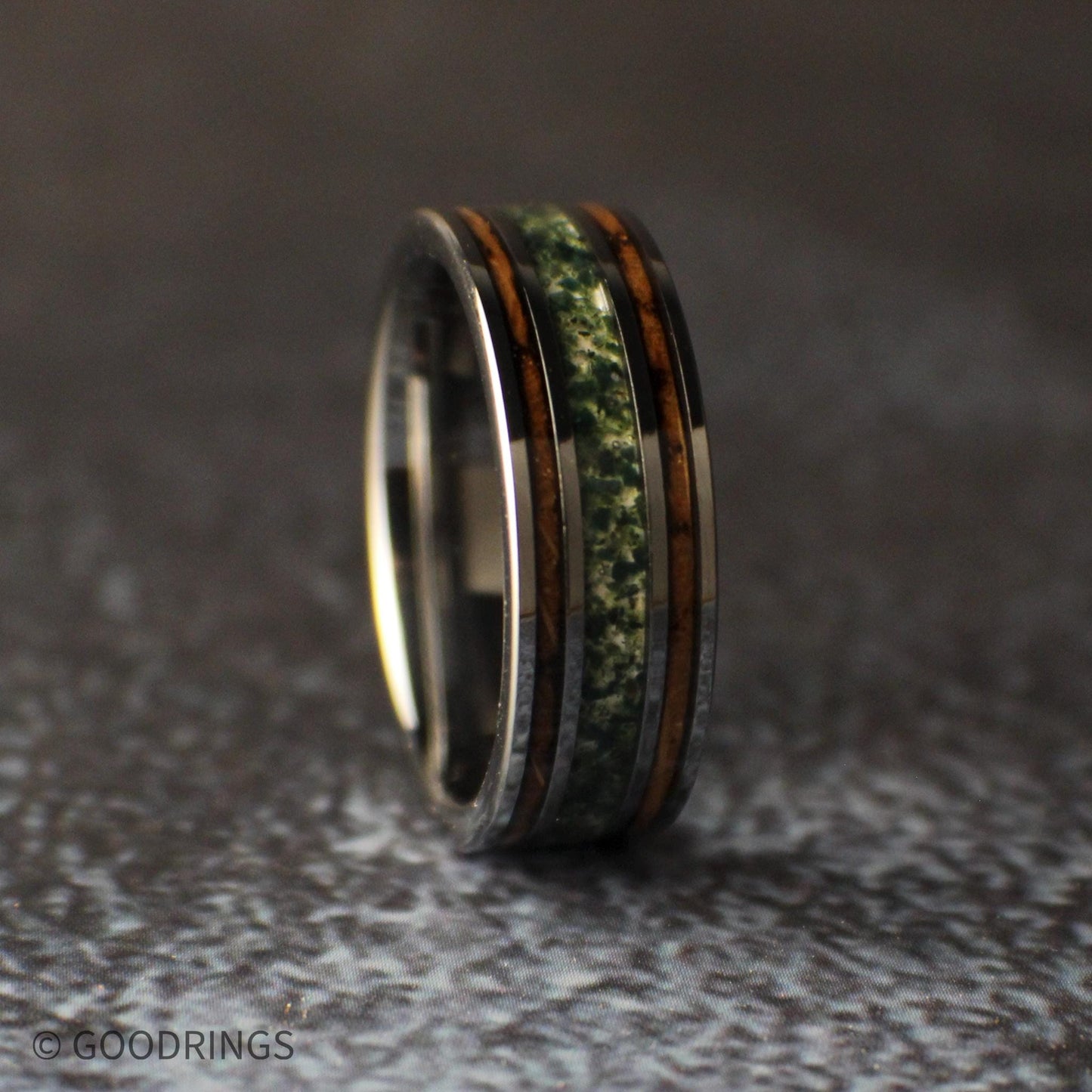 Close angled view of the moss agate and whiskey barrel wood tungsten ring leaning against a gray stone on a dark textured background.
