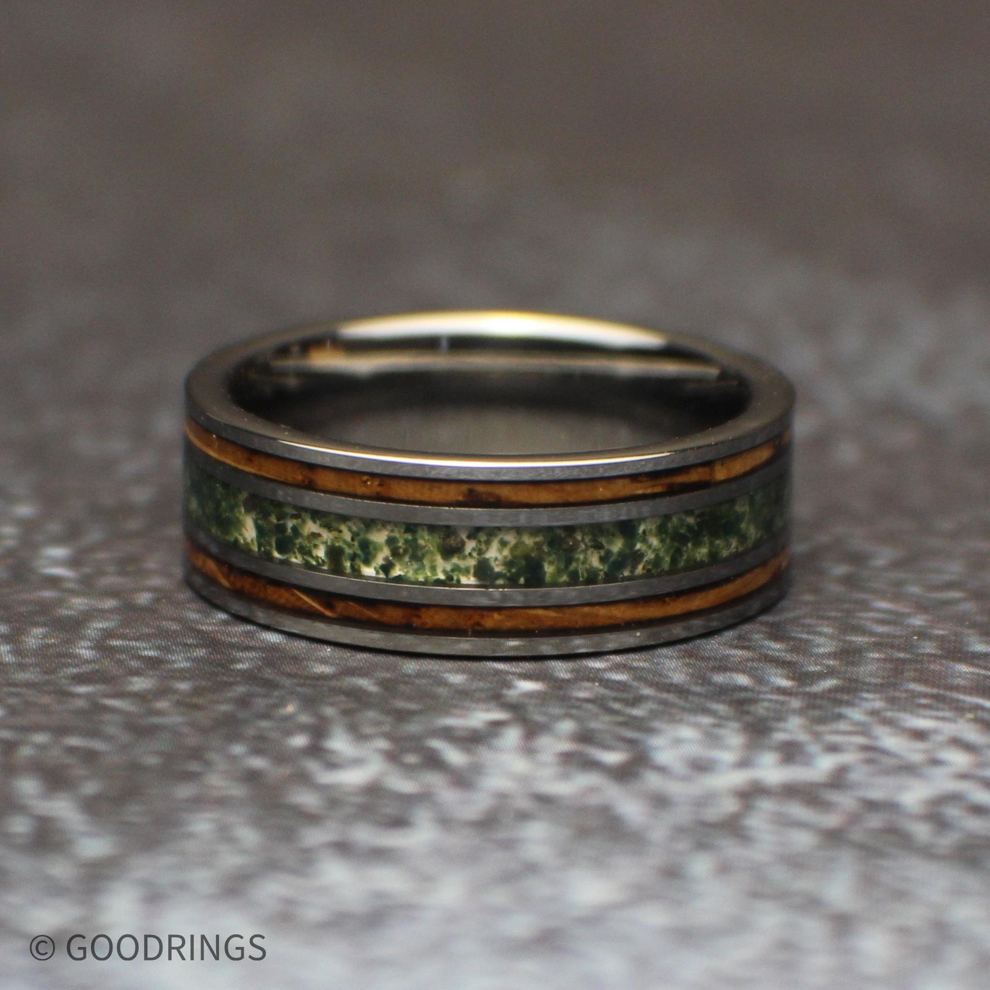 Low front view of the tungsten ring lying on its side, highlighting the mottled green moss agate center and parallel brown wood inlays.
