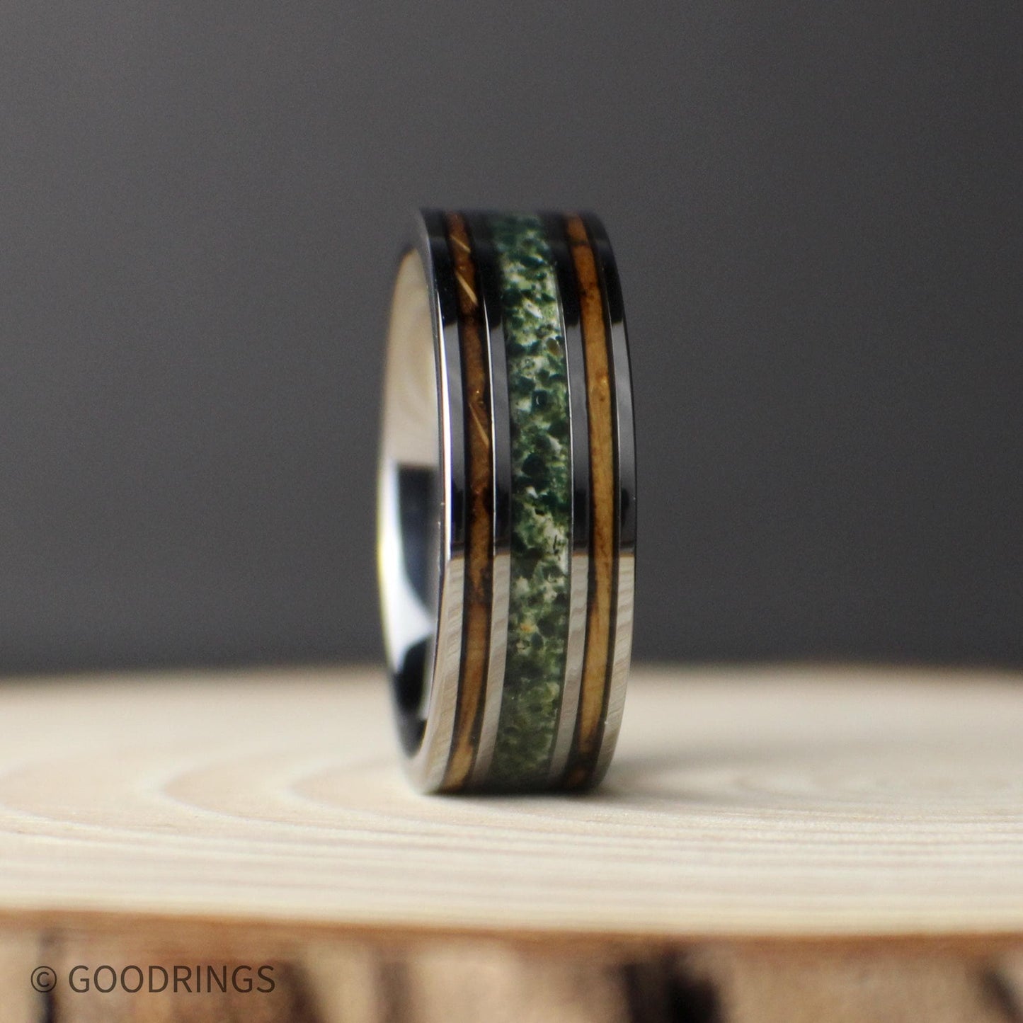 Three-quarter view of a polished gunmetal tungsten ring standing upright, with a green moss agate center inlay and narrow whiskey barrel wood inlays on a dark textured surface.