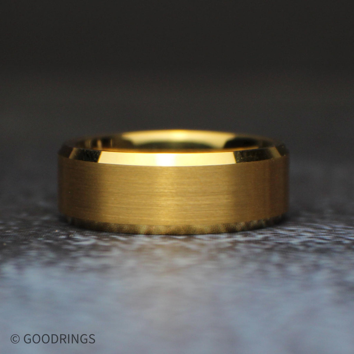 Three-quarter view of a men’s brushed gold tungsten ring standing on a dark textured surface, with polished beveled edges reflecting the light.