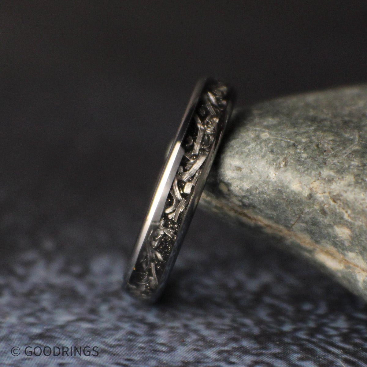 Close side view of the 4mm meteorite tungsten ring lying on a gray stone, showing the varied shapes and textures of the silver shavings.