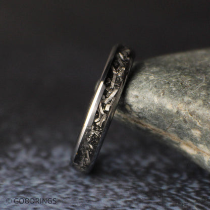 Close side view of the 4mm meteorite tungsten ring lying on a gray stone, showing the varied shapes and textures of the silver shavings.