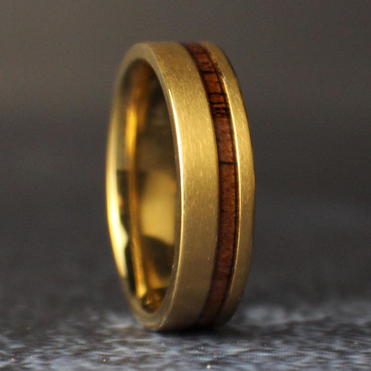 Close three-quarter view of a 6mm brushed gold-plated titanium ring standing upright, showing its narrow offset koa wood inlay and polished interior.