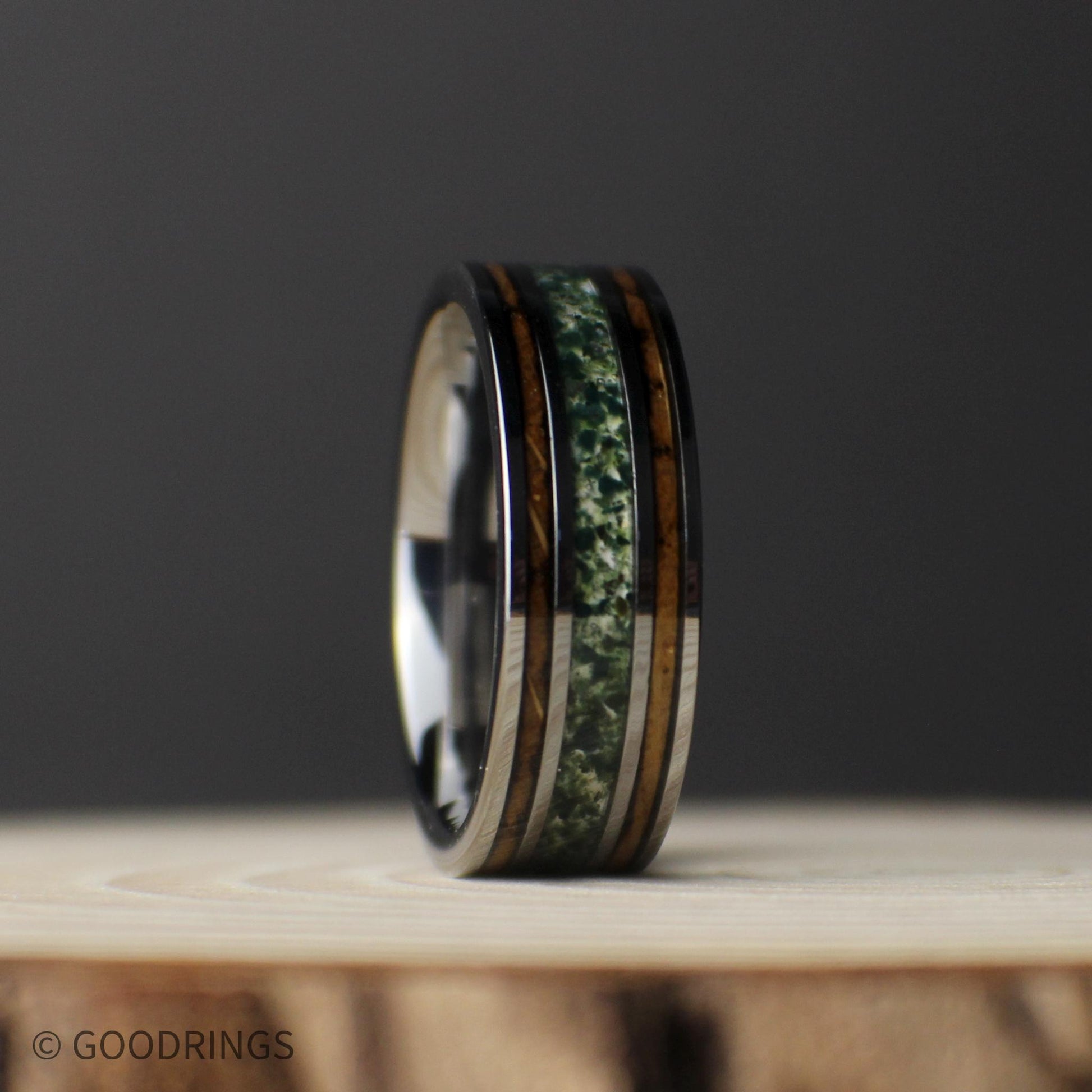 Elevated front view of the gunmetal tungsten band lying flat, showing its polished comfort-fit interior, green moss agate center, and whiskey barrel wood inlays.