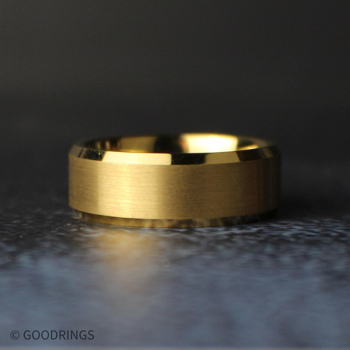 Low angled view of the gold tungsten ring lying on its side, showing the wide brushed center, reflective beveled edges, and polished interior.