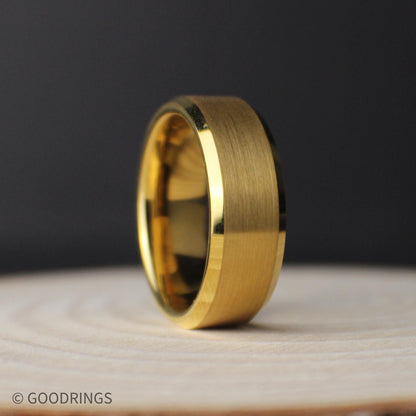 Eight-millimeter brushed gold tungsten band standing upright on a light wood surface, displaying its polished bevels and smooth gold interior.