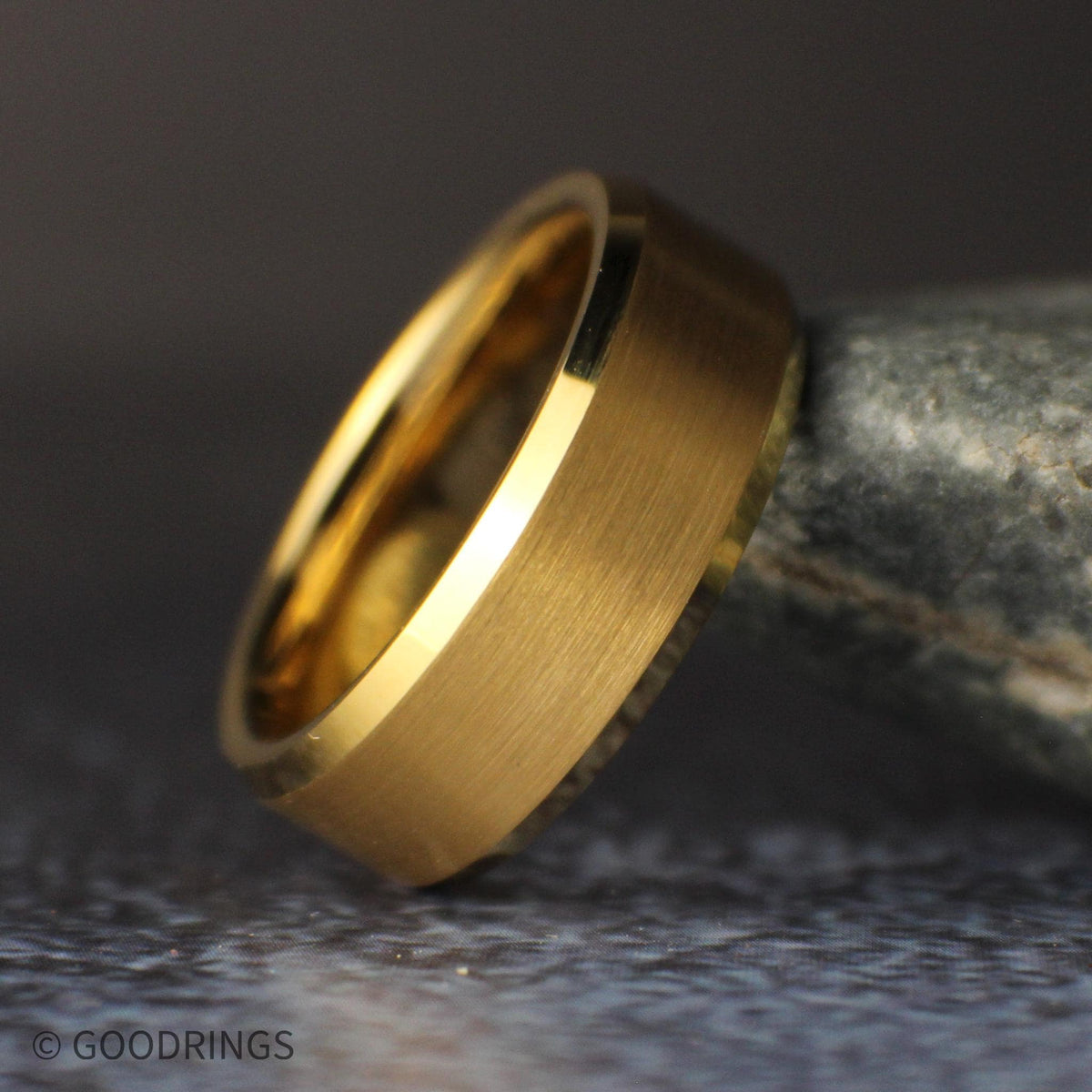 Close side view of the brushed gold tungsten wedding band leaning against a gray stone, highlighting the satin center and shiny beveled edges.