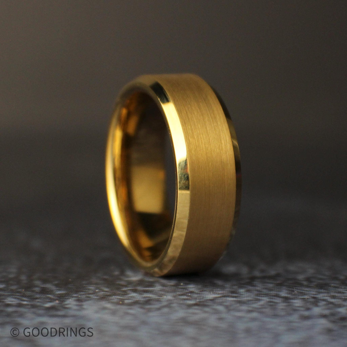 Close angled view of an 8mm gold tungsten wedding band standing upright, showing its brushed center, polished beveled edges, and gold comfort-fit interior.