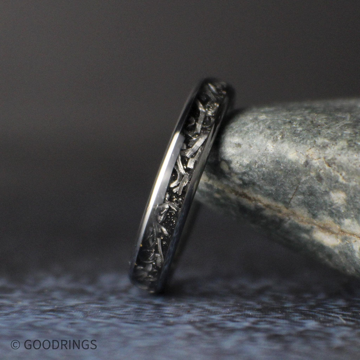 Close angled view of a slim 4mm tungsten ring leaning against a gray stone, showing silver meteorite shavings set in a dark inlay.