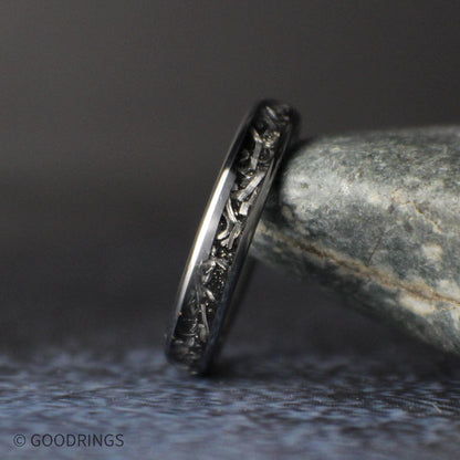 Close angled view of a slim 4mm tungsten ring leaning against a gray stone, showing silver meteorite shavings set in a dark inlay.