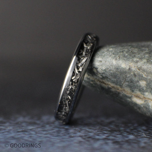 Close angled view of a slim 4mm tungsten ring leaning against a gray stone, showing silver meteorite shavings set in a dark inlay.