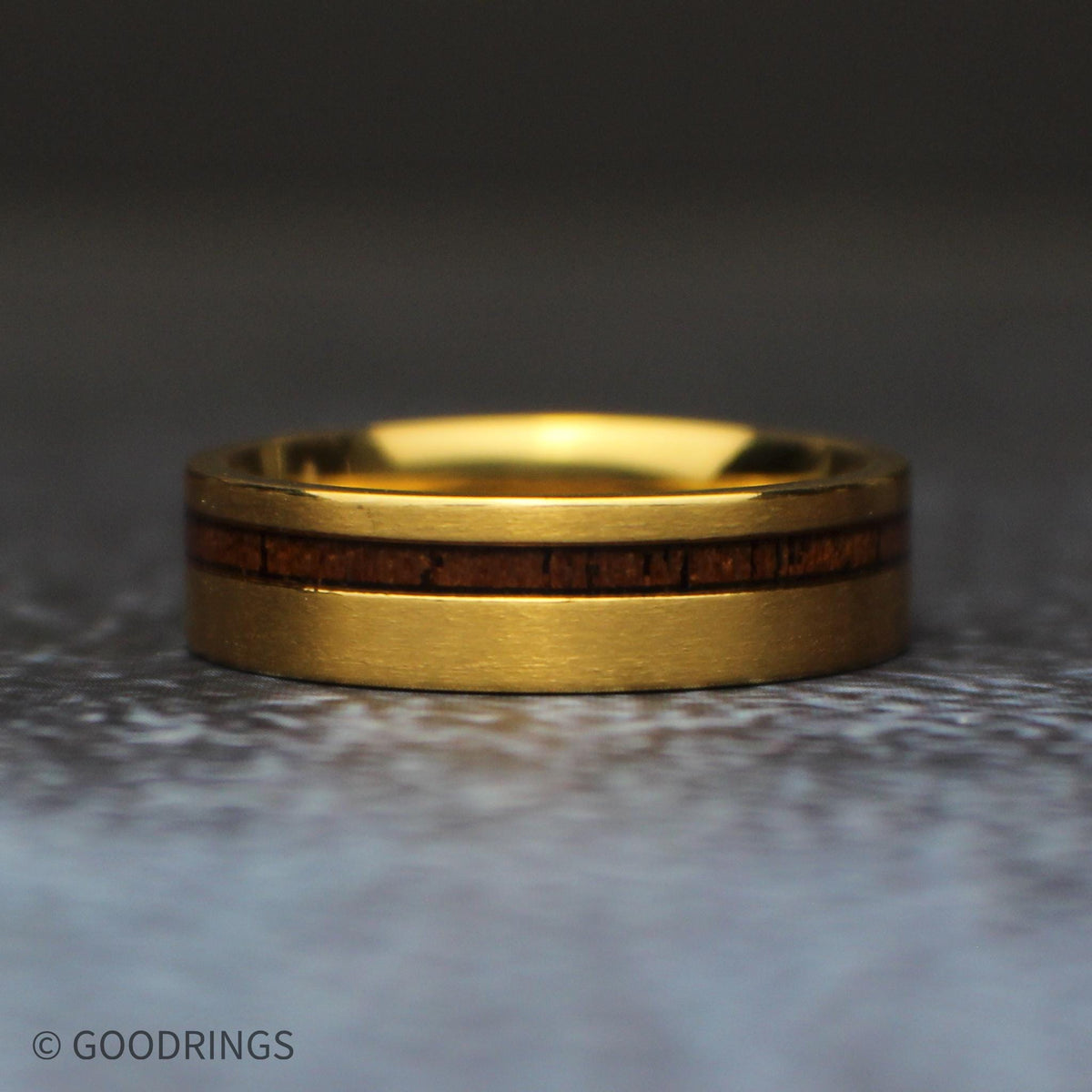 Straight front view of the 6mm gold titanium wedding band lying horizontally, with a narrow koa wood inlay positioned above the center of the brushed band.