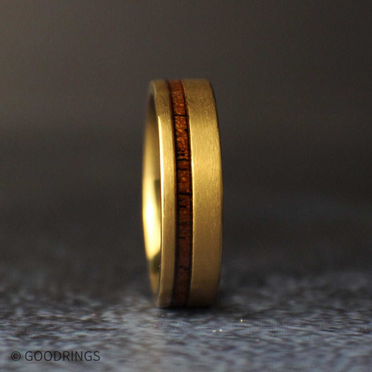 Low angled view of the gold-plated titanium band lying on a gray stone, highlighting its brushed finish, polished interior, and offset koa wood stripe.