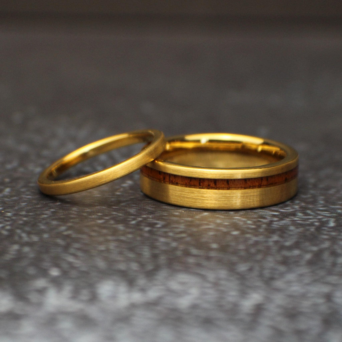 Matching gold tungsten wedding rings lying together, with a slim 2mm brushed band overlapping a wider 6mm band featuring an offset koa wood inlay.