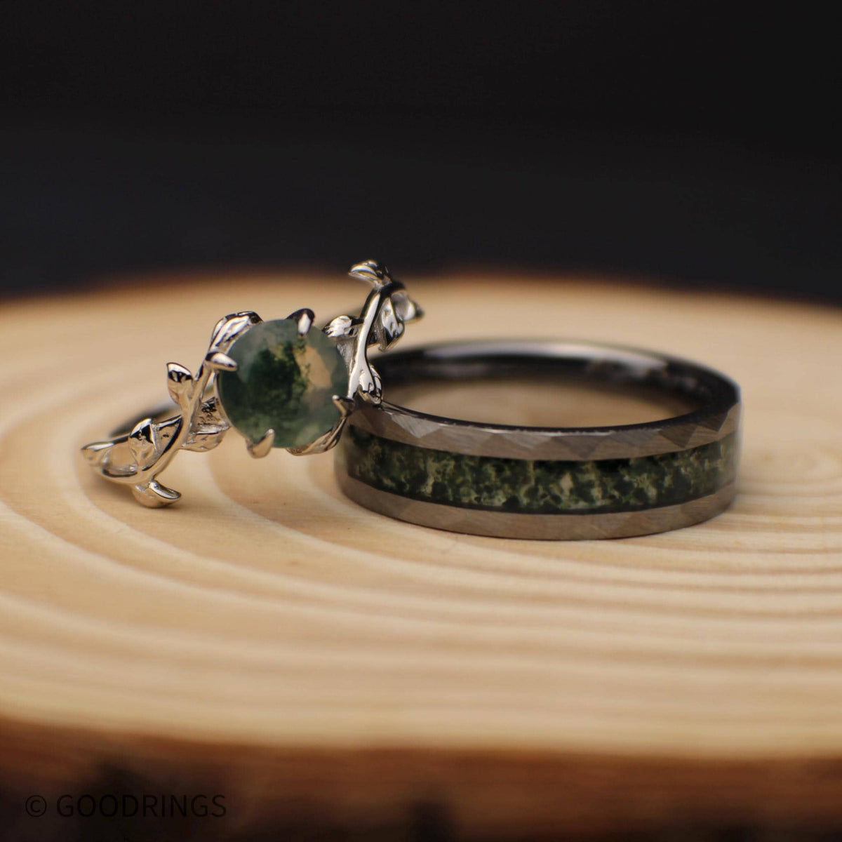 Picture of a female moss agate engagement ring leaning on a hammered mens tungsten wedding band inlay with moss agate.
